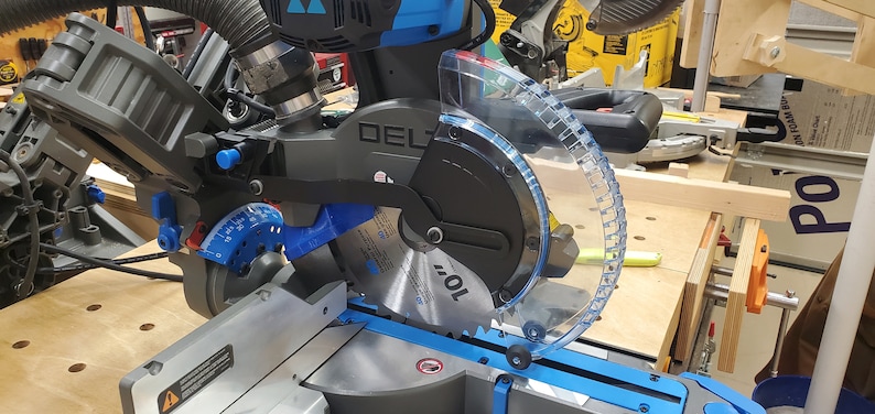 May include: A close-up of a black and blue Delta power miter saw with a clear plastic blade guard. The saw is on a wooden table and the blade is visible.