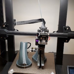 May include: A 3D printer with a black frame and a white filament spool. The printer is printing two gray objects on a black platform. The printer has a touch screen display with the text "Creality" and a logo.
