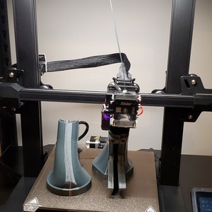 May include: A 3D printer with a black frame and a white filament spool. The printer is printing two gray objects on a black platform. The printer has a touch screen display with the text "Creality" and a logo.