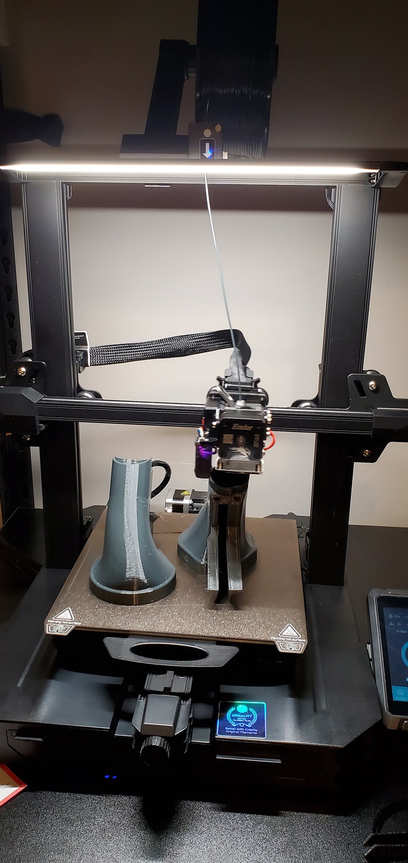 May include: A 3D printer with a black frame and a white filament spool. The printer is printing two gray objects on a black platform. The printer has a touch screen display with the text "Creality" and a logo.