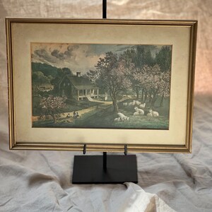 May include: A framed landscape print featuring a house with a porch, a dirt path, and a group of sheep grazing under flowering trees. The artwork is enclosed in a gold-toned frame with a cream-coloured mat, displayed on a black metal stand.