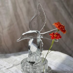 May include: A clear glass sculpture shaped like a bird in flight, featuring a circular base with a small opening for a flower. Two vibrant orange flowers with green stems are inserted into the base, positioned next to the glass bird figure.