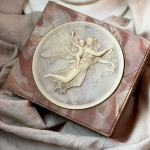 May include: A square box with a marble-effect finish in pink and white, featuring a circular, off-white relief carving. The carving depicts a winged figure and a child. The box is a decorative item.