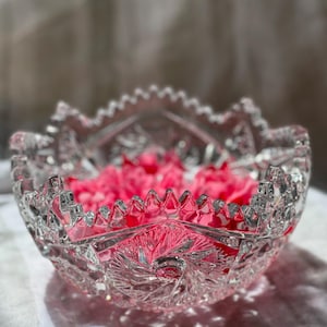 May include: A decorative cut-glass bowl filled with pink flower petals. The bowl has a starburst design in the centre and a scalloped edge. The light reflects off the glass, creating shadows on the white surface beneath.