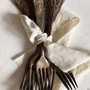 May include: A bundle of antique silver forks tied with a cream-colored fabric. The forks have ornate, detailed handles and are arranged in a fan shape. The fabric is knotted around the fork tines, creating a rustic and elegant presentation.