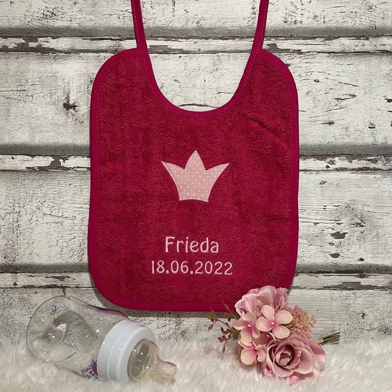 May include: A pink and white baby bib with a crown design and the name "Frieda" and the date "18.06.2022" embroidered on it.