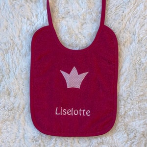 May include: A pink baby bib with a white crown and the name "Liselotte" embroidered on it.