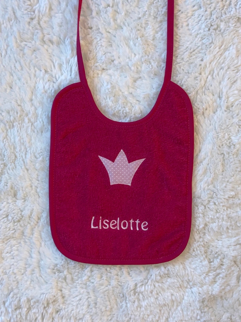 May include: A pink baby bib with a white crown and the name "Liselotte" embroidered on it.