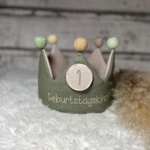 Könnte beinhalten: Eine grüne Filzkrone mit einem weißen Kreis, auf dem die Zahl "1" und der Text "Geburtstagskind" gestickt sind. Die Krone ist mit bunten Filzkugeln verziert.