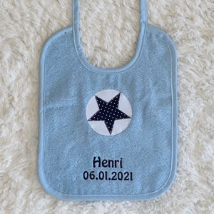 May include: Light blue baby bib with a white circle and a black star with white polka dots. The bib has the name Henri and the date 06.01.2021 embroidered on it.