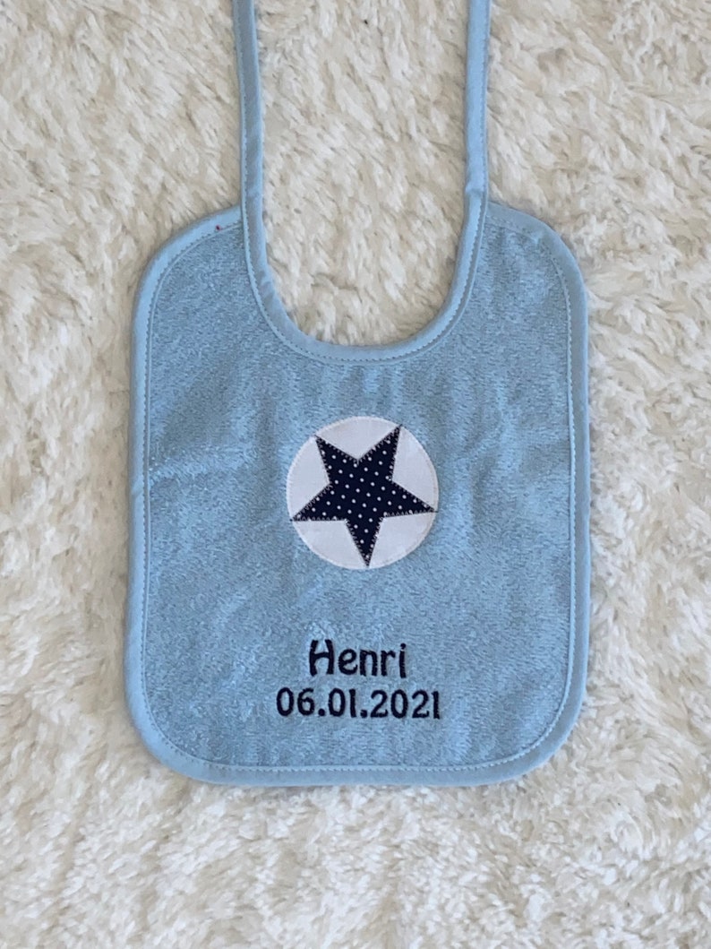 May include: Light blue baby bib with a white circle and a black star with white polka dots. The bib has the name Henri and the date 06.01.2021 embroidered on it.
