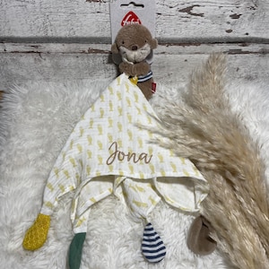 Könnte beinhalten: Eine weiße Musselin-Babydecke mit gelben Fischen bedruckt. Die Decke hat einen grünen, gelben und blauen Saum. Die Decke ist mit dem Namen "Jona" personalisiert. Ein braunes Plüsch-Seeotter-Spielzeug ist an der Decke befestigt.