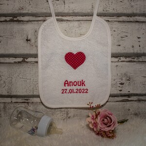 May include: White cotton baby bib with a red heart with white polka dots and the name "Anouk" embroidered in red with the date "27.01.2022" underneath.