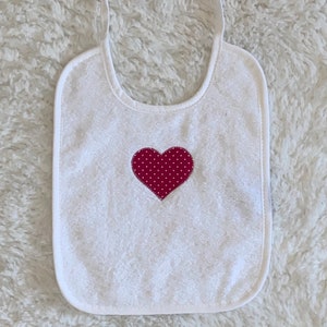 May include: White terry cloth baby bib with adjustable straps and a red heart with white polka dots in the centre.