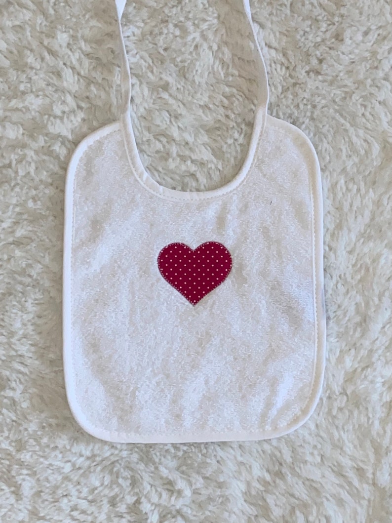 May include: White terry cloth baby bib with adjustable straps and a red heart with white polka dots in the centre.