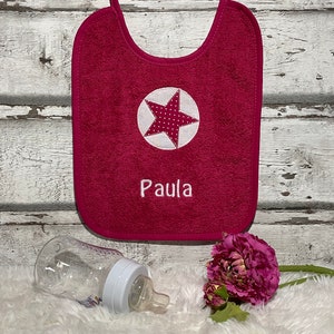 May include: A pink terry cloth baby bib with a white star and the name "Paula" embroidered on it. The bib has a white polka dot star in the centre and is hanging on a white wooden background.