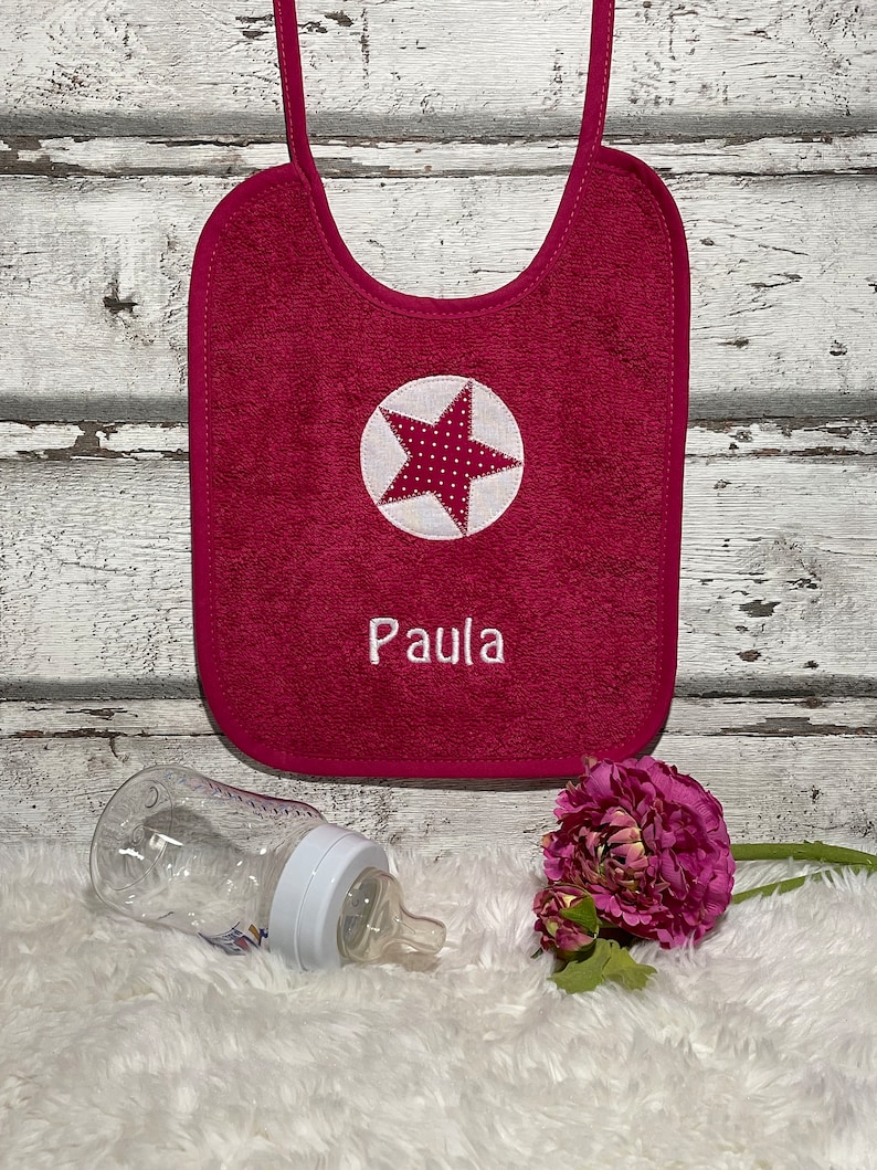 May include: A pink terry cloth baby bib with a white star and the name "Paula" embroidered on it. The bib has a white polka dot star in the centre and is hanging on a white wooden background.