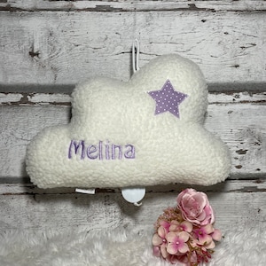 Könnte beinhalten: Ein weißes, wolkenförmiges Plüschtier mit dem Namen "Melina" in Lila gestickt. Ein lila Stern mit weißen Punkten ist auf die Wolke genäht. Das Spielzeug hat eine Aufhängeschlaufe und eine kleine Spieluhr. Ein Strauß rosa Blumen befindet sich im Vordergrund.
