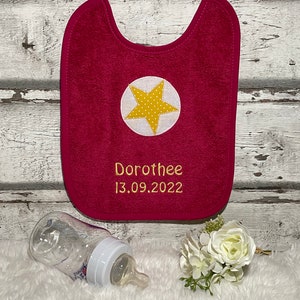 May include: A pink baby bib with a yellow star and the name "Dorothee" and the date "13.09.2022" embroidered on it.