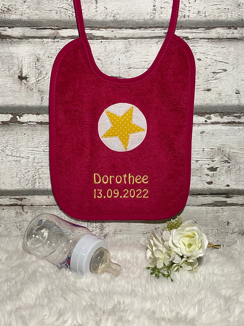 May include: A pink baby bib with a yellow star and the name "Dorothee" and the date "13.09.2022" embroidered on it.