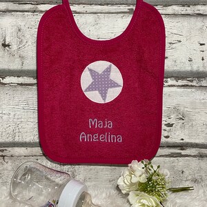 May include: A pink baby bib with a white star with purple polka dots and the name "Maja Angelina" embroidered on it.