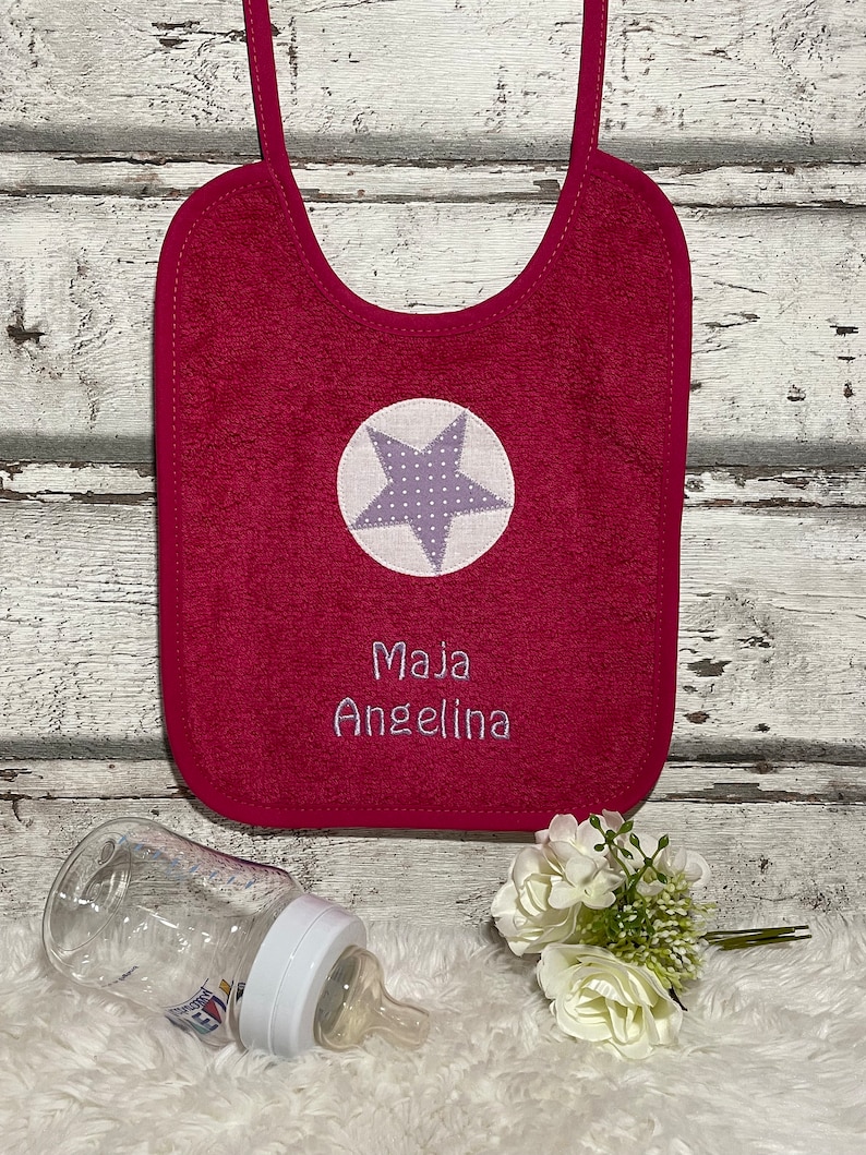 May include: A pink baby bib with a white star with purple polka dots and the name "Maja Angelina" embroidered on it.