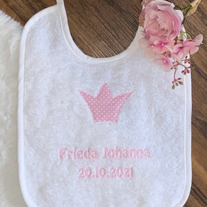 May include: White baby bib with a pink embroidered crown and the text "Frieda Johanna 20.10.2021".