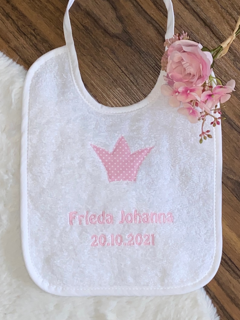 May include: White baby bib with a pink embroidered crown and the text "Frieda Johanna 20.10.2021".