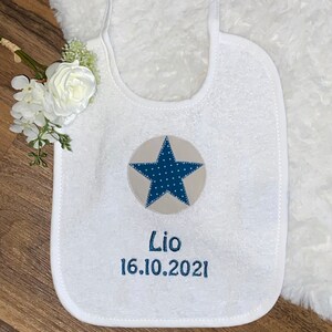 May include: White baby bib with a blue and beige star and the text "Lio 16.10.2021" embroidered on it.