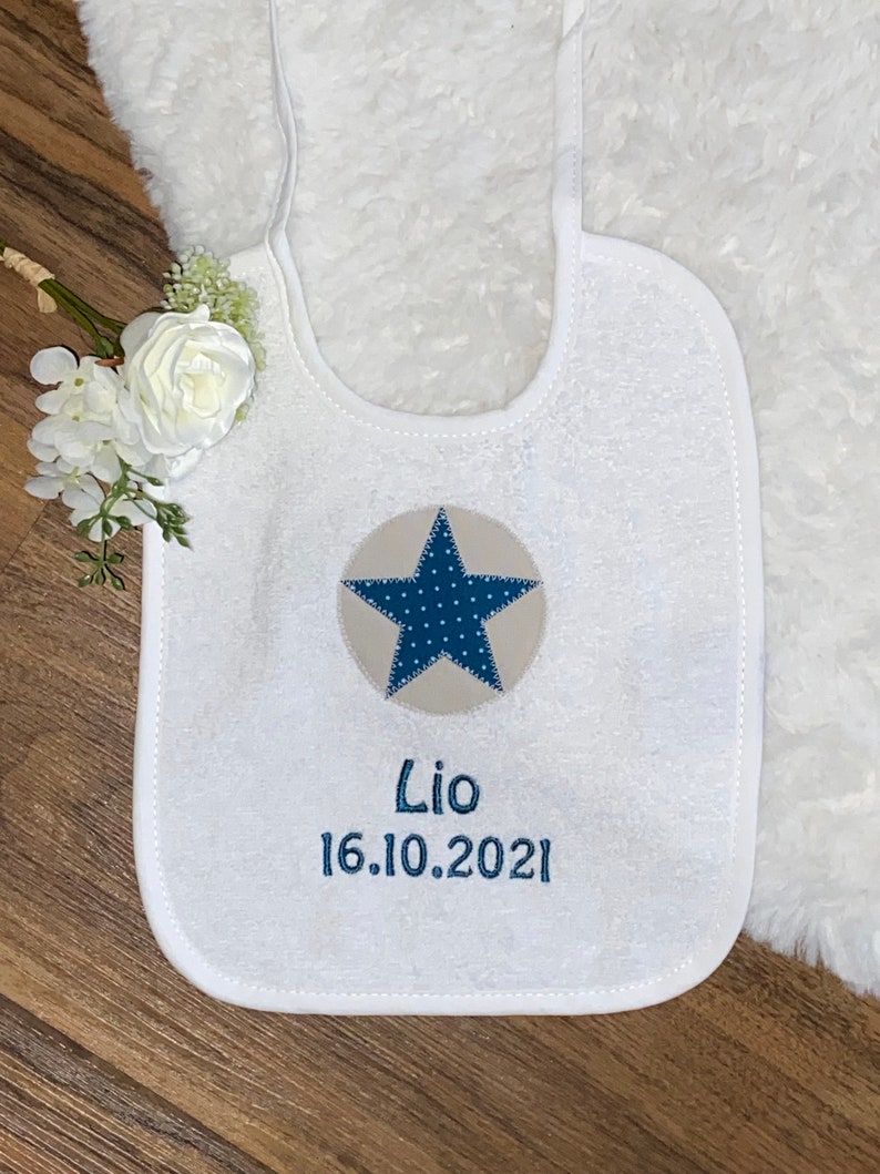 May include: White baby bib with a blue and beige star and the text "Lio 16.10.2021" embroidered on it.