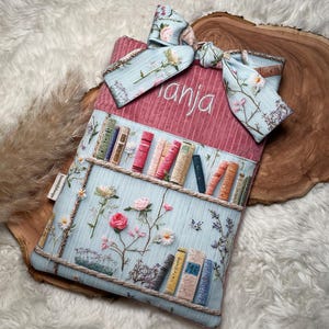 Könnte beinhalten: Eine rechteckige Buchhülle aus Stoff mit einem oberen Teil aus altrosa Kord, bestickt mit dem Namen "Anja", und einem hellblauen unteren Teil, bedruckt mit einem Bücherregal, bunten Buchrücken und Blumenmustern, abgerundet mit einer passenden Schleife.