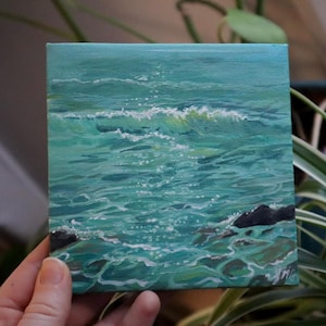 May include: Square canvas painting depicting ocean waves in shades of teal and turquoise. White foam crests the waves, and dark rocks are visible near the bottom. The artwork is held by a hand, with green foliage in the background.