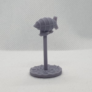 Puede incluir: Una miniatura gris de un dirigible impresa en 3D sobre un soporte. El dirigible tiene una cesta y una hélice. El soporte es una simple base redonda.
