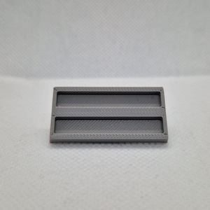 Puede incluir: Un objeto rectangular gris impreso en 3D con dos secciones empotradas y una superficie texturizada. El objeto tiene una serie de pequeños agujeros a lo largo de los bordes superior e inferior. El diseño es simple y funcional.