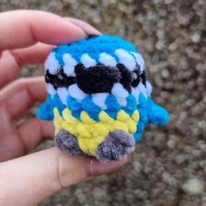 Może przedstawiać: Ręcznie robiona figurka ptaka na szydełku. Ptak ma niebieską, białą i czarną głowę, żółtą pierś i szare stopy. Mały, pluszowy ptak jest trzymany w dłoni na rozmytym tle.