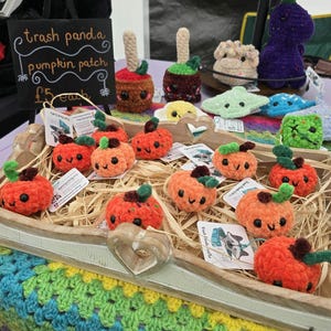 Op de afbeelding: Een houten dienblad gevuld met kleine, oranje, gehaakte pompoenfiguren, elk met een groen steeltje en een lachend gezichtje. Een zwart bordje met witte tekst leest trash panda pumpkin patch £5 each. Andere gehaakte items zijn op de achtergrond zichtbaar.