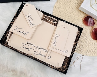 Puede incluir: Una caja de regalo negra con una inscripción dorada que dice "A Lifetime of Adventures Kushal & Krishali". La caja contiene un conjunto de accesorios de viaje, incluyendo un portapasaporte, etiquetas de equipaje y un portadocumentos de viaje. Los accesorios están hechos de cuero beige y tienen los nombres "Kushal" y "Krishali" escritos en ellos.
