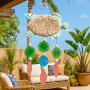 May include: A wooden turtle wind chime with a light brown shell and turquoise detailing. The chime features green, blue, and pink decorative elements. It hangs from a natural rope, suitable for outdoor decoration.