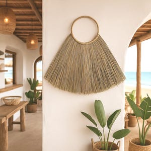 May include: A natural fibre wall hanging with a fan-like shape. The decorative piece features a circular top and a wide, textured base made of light brown strands. It hangs on a white wall, adding a touch of bohemian style to the space.