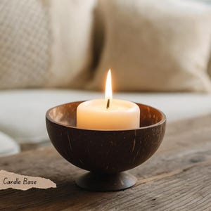 May include: A burning ivory candle is placed in a dark brown coconut shell candle holder. The candle holder has a round base and a bowl-shaped top. The text "Candle Base" is visible in the lower left corner. The background is blurred.
