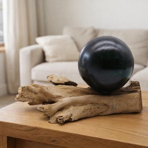 May include: A dark, spherical ornament with green and blue highlights sits on a piece of driftwood, displayed on a wooden table. The driftwood has a natural, weathered appearance. The decor has a rustic, natural theme.
