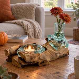 May include: A decorative piece featuring a natural piece of driftwood with a molten glass bowl and a molten glass vase fused to the wood. The glass bowl holds a lit tea light candle, and the glass vase contains two orange chrysanthemum flowers.