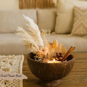 May include: A decorative dried flower bowl made from a coconut shell. The bowl contains a lit candle, dried flowers, a pine cone, and cinnamon sticks. The text "Dried Flower Bowl" is visible on a label.