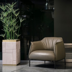 Può includere: Una poltrona in pelle marrone chiaro con un design plissettato si trova su un pavimento grigio scuro di fronte a una grande pianta in vaso. La pianta è in un vaso rosa e oro con un design geometrico.