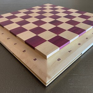 Puede incluir: Un tablero de ajedrez de madera con un acabado en madera natural y morado. El tablero est&aacute; doblado por la mitad, mostrando los n&uacute;meros y las letras de cada casilla.