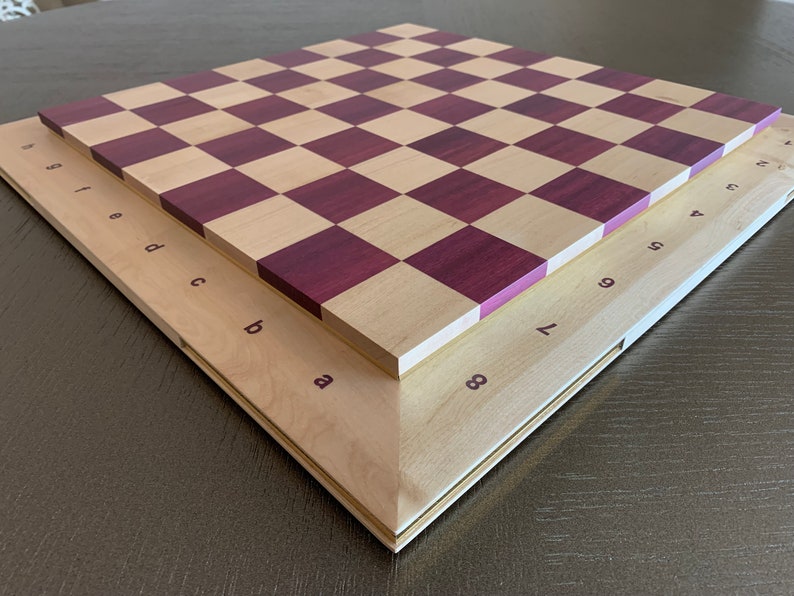 Puede incluir: Un tablero de ajedrez de madera con un acabado en madera natural y morado. El tablero est&aacute; doblado por la mitad, mostrando los n&uacute;meros y las letras de cada casilla.