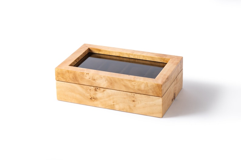 May include: A wooden watch box with a glass top. The box has a light brown wood grain and is made of two pieces. The top piece has a glass window that shows the interior of the box. The box is empty.