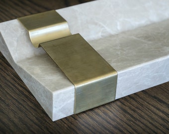 May include: A rectangular tray made of light gray marble with two brass accents. The tray has a slightly curved edge and a smooth surface.