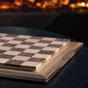 May include: A wooden chessboard with a dark brown and light wood checkered pattern. The board is closed and sitting on a black surface.