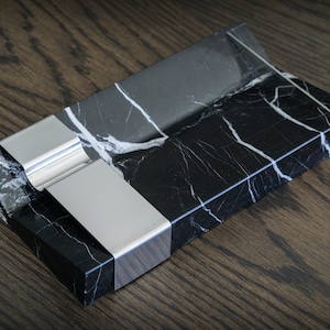 以下が含まれることがあります： 黒と白の大理石のトレイで、銀色の金属製のインサートが付いています。トレイは長方形で、わずかに湾曲した縁があります。